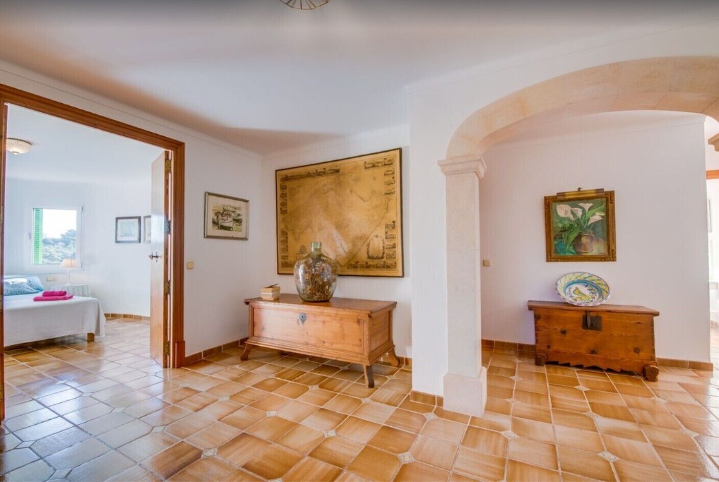 A room with a bed, a wooden dresser, a map on the wall, and a view through an open door.