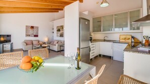 A modern kitchen with white cabinets, a built-in oven, and a dining area with a glass table and wicker chairs. - Seafront Penthouse “Es Port” with Sea View, Mountain View, Wi-Fi, A/C & Balcony (Santanyí)