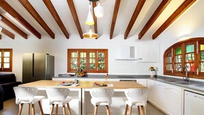 A modern kitchen with wooden beams, a central island with bar stools, a refrigerator, and a window with a view. - Finca “Jaume Romona” with Mountain View, Pool, Wi-Fi, Air Conditioning, Terrace & Garden (Pollensa)