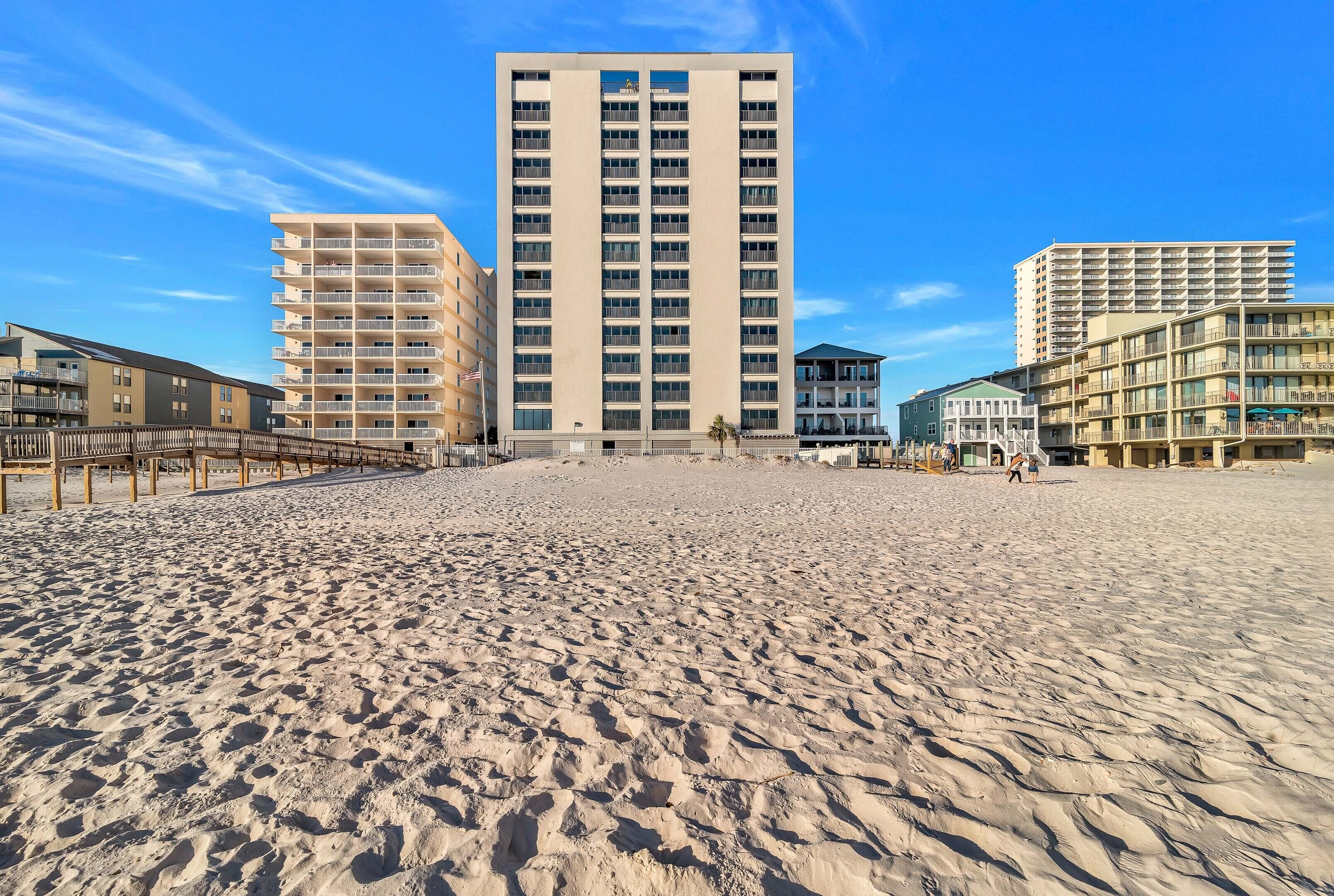 Una playa con un edificio alto en el centro, un paseo marítimo de madera a la izquierda y un edificio más pequeño a la derecha.