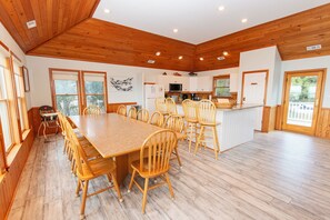 A spacious dining area with a long wooden table and chairs, a kitchen with white cabinets, and a view of the outdoors through large windows. - Soundfront, Sound views, Private Pool, Hot Tub! 509, Friday Harbor (Corolla)
