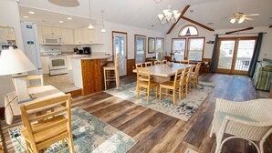 A spacious kitchen with wooden cabinets, a dining area with a large table and chairs, and a cozy seating area with a wicker chair. - Oceanside, Dogs Welcome, Private Pool, Hot Tub, Close to Beach Access, OL626, Celtic Dunes (Corolla)