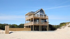 Une maison à plusieurs étages située en bord de mer, dotée d’une terrasse et d’un balcon en bois, et entourée de sable et de végétation.