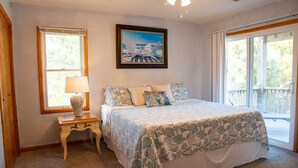 A bedroom with a bed, a nightstand, a lamp, a framed picture, and a window with a view of the outdoors.