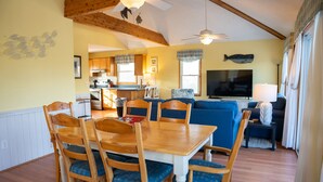 A dining area with a wooden table and chairs, a kitchen in the background, and a living area with a sofa and TV.
