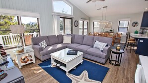 Ein geräumiges Wohnzimmer mit einem großen, grauen Ecksofa, einem Couchtisch und einem Teppich mit Seestern-Muster. Es gibt einen Essbereich mit Holzhöhlen und einem Tisch, sowie eine Küche mit Spüle und Schränken.