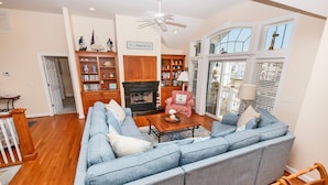 A living room with a blue sofa, wooden coffee table, and a fireplace. There are bookshelves and a chandelier. - Oceanside, Private Pool, Community Amenities, Hot Tub, VOH94, Knot A Problem (Corolla)