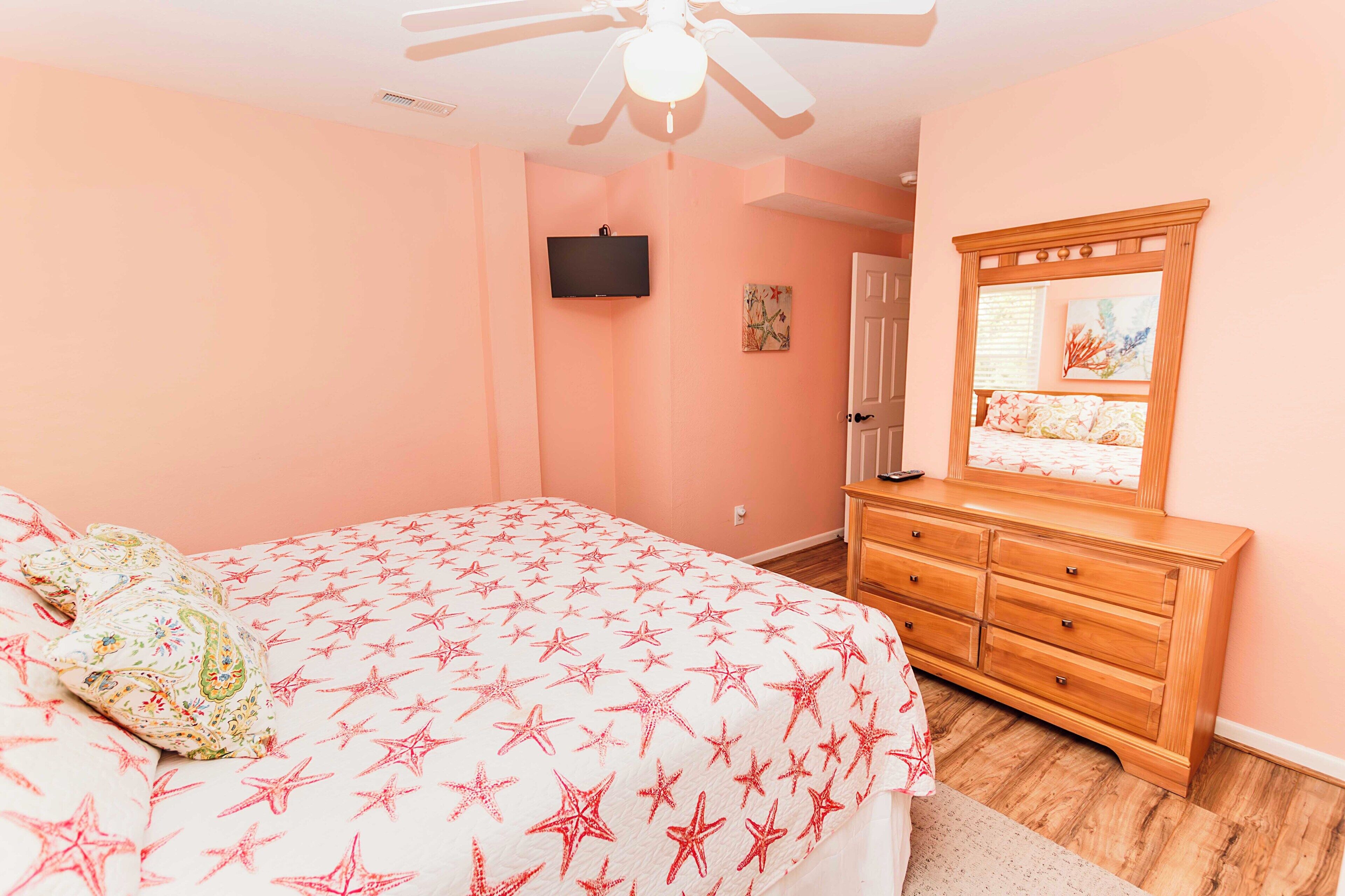 A bedroom with a bed, a dresser, a ceiling fan, a TV, and a door.