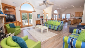 A spacious living room with a large sofa, green armchairs, and a television. There is a kitchen area with wooden cabinets and a dining table. - Oceanside, Heated Pool, Poolside Pavilion, Hot Tub, 4, Relax Mom (Corolla)