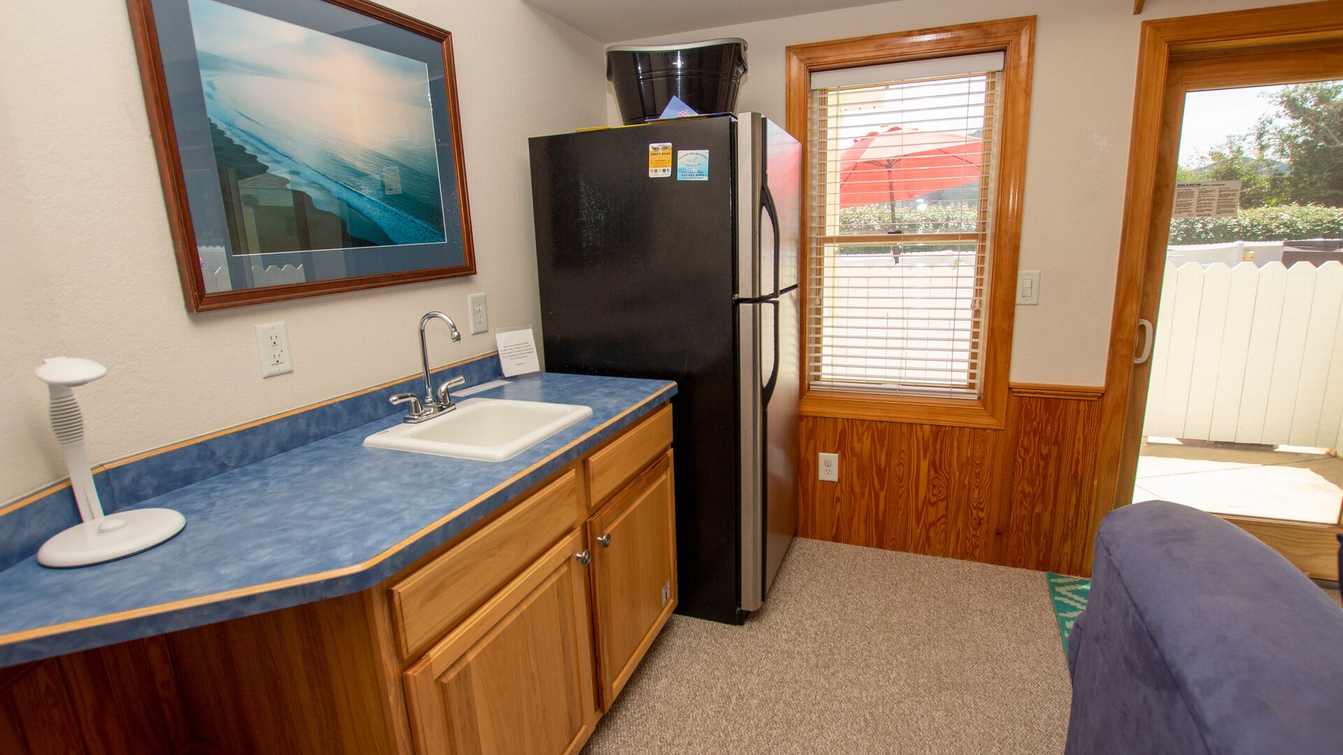 A compact kitchen with a sink, a black refrigerator, wooden cabinets, and a window with blinds.