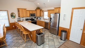 A kitchen with wooden cabinets, a central island with bar stools, stainless steel appliances, and a trash can. - Oceanside, Heated Pool, Poolside Pavilion, Hot Tub, 4, Relax Mom (Corolla)