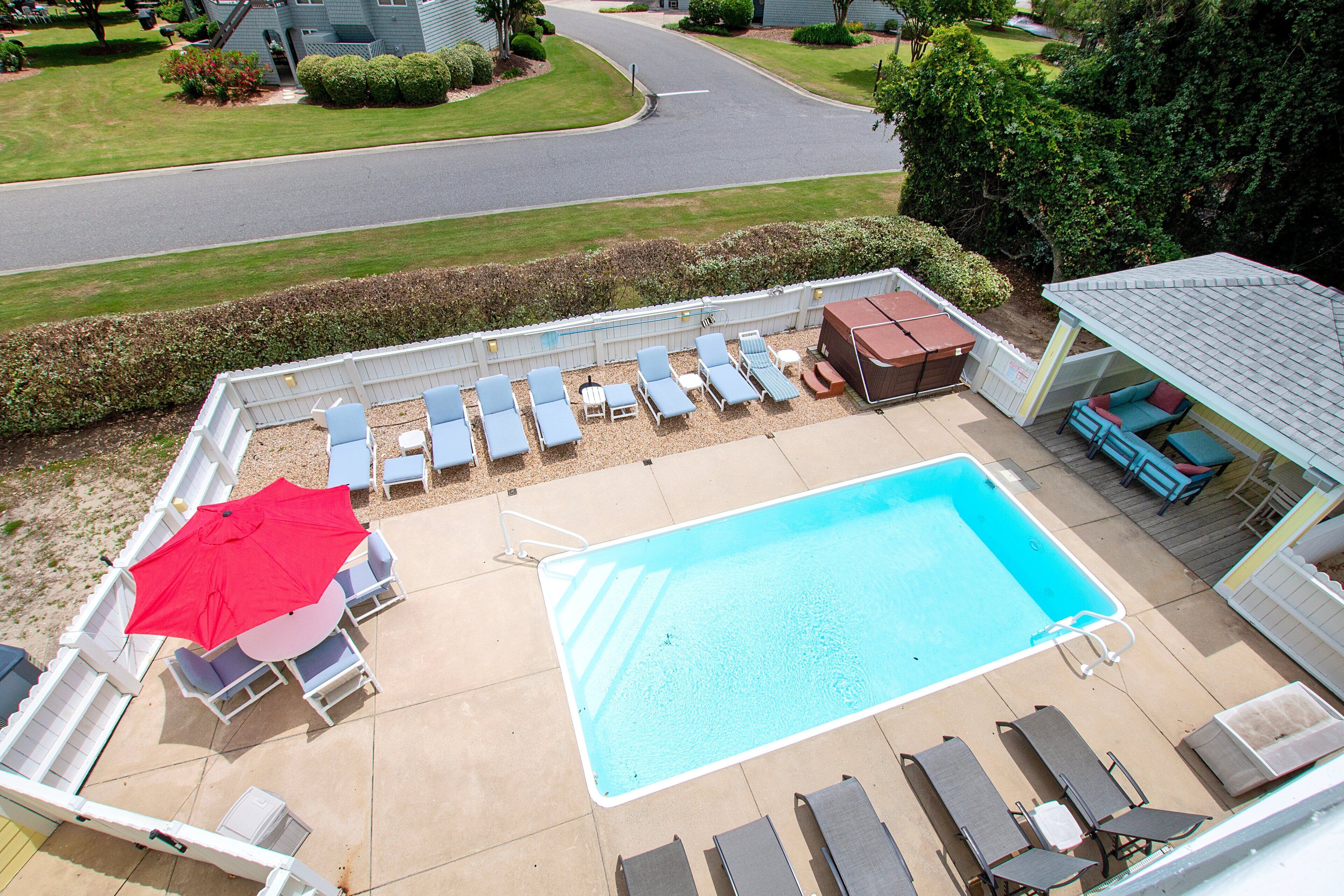 A rectangular swimming pool with lounge chairs and an umbrella, surrounded by a white fence and a covered seating area.
