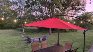 Un pati amb una taula i cadires de fusta, un parasol vermell i llums penjades.