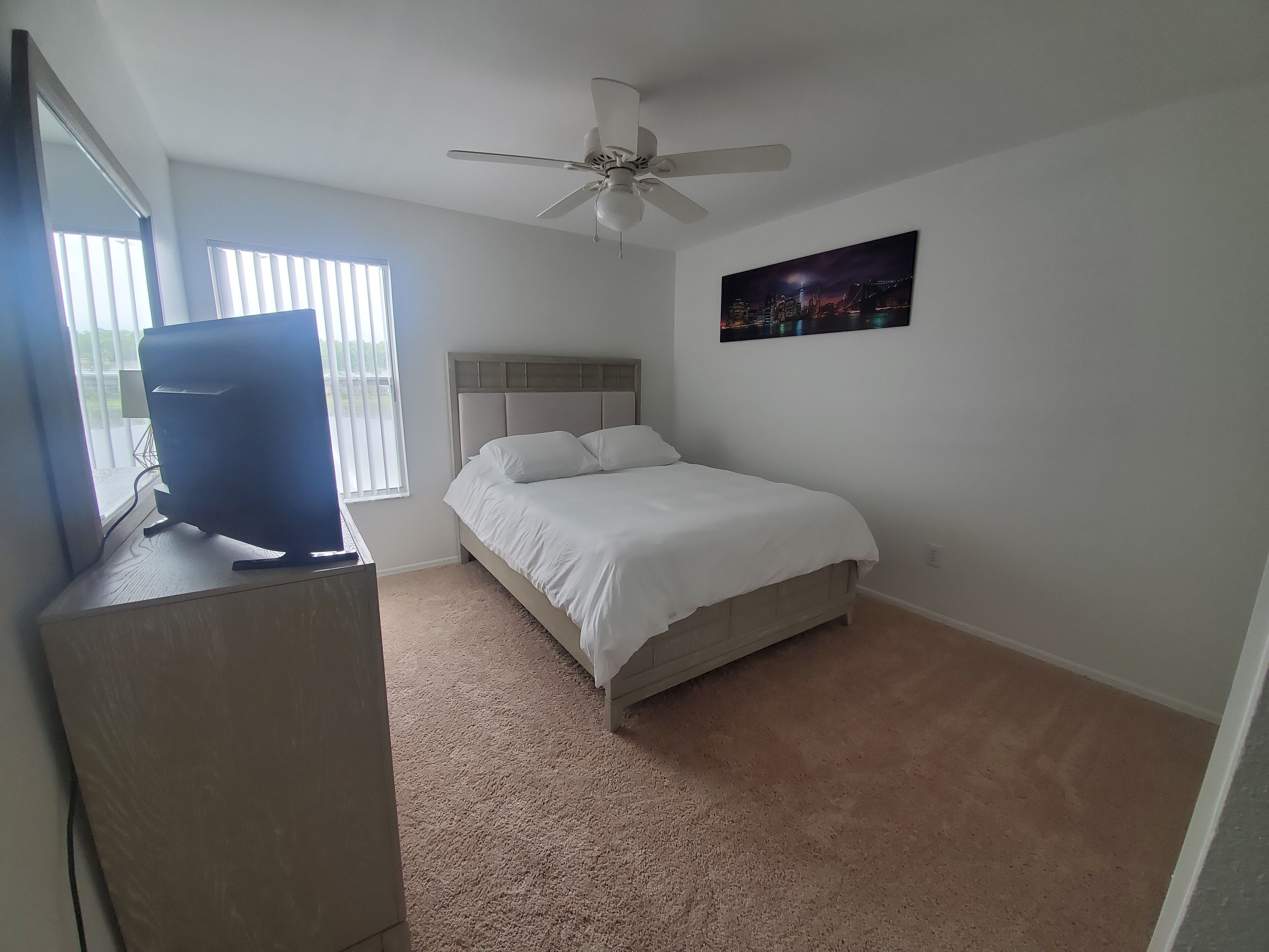 A bedroom with a bed, a ceiling fan, a TV on a stand, and a cityscape painting on the wall.