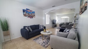 Sala de estar moderna com um sofá azul escuro, uma poltrona cinza e uma mesa de centro. Há uma janela grande com persianas, um quadro na parede e uma planta numa cesta de fibra.