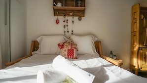A neatly made bed with white linens, a decorative pillow, and a wooden headboard. A wooden shelf above the bed holds a few items.