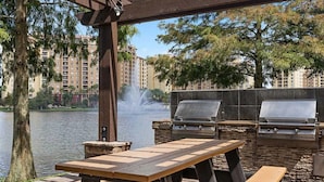 An outdoor patio with a grill and a table, overlooking a body of water and a building complex.