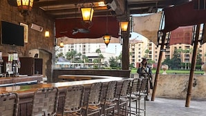 A bar area with a stone wall, a flat-screen TV, and a variety of liquor bottles. There are metal chairs and a wooden bar counter.