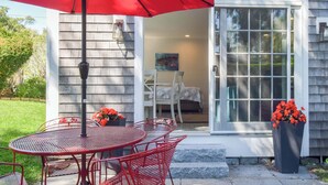 Une terrasse avec un parasol rouge, une table en métal et des chaises rouges. On y trouve deux pots de fleurs contenant des fleurs rouges.