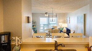 A modern kitchen with a sink, a coffee maker, and a dining area with white chairs and a table. - Turtle Dunes #1854 - Ocean Views (amelia island)