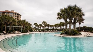 A large, clear swimming pool with a curved edge, surrounded by palm trees and lounge chairs. - Turtle Dunes #1854 - Ocean Views (amelia island)