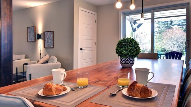 Une table en bois, recouverte de sets de table, est dressée pour le petit-déjeuner avec deux croissants, deux verres de jus d’orange et deux tasses blanches.