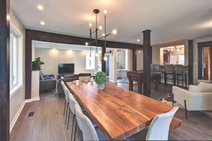A spacious living area with a large wooden dining table, white chairs, a sofa, and a television.