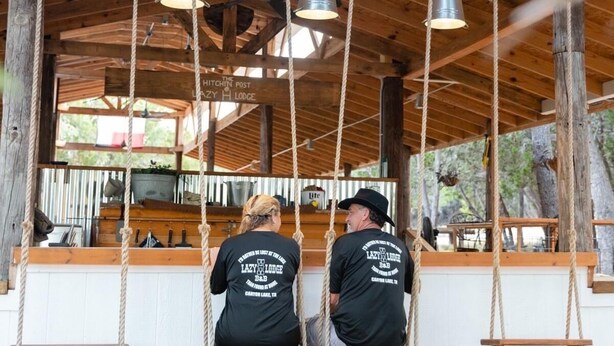 Two people on a swing with a rustic wooden structure and a sign reading "Lazy Lodge."