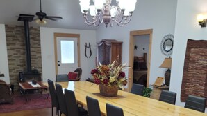 A dining area with a long wooden table, black chairs, and a chandelier. There is a fireplace and a ceiling fan in the background.