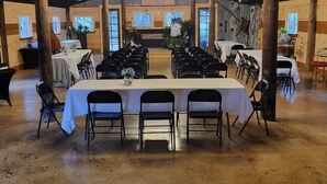 A large hall with a high ceiling and wooden beams, featuring a central stage with a chandelier, surrounded by rows of tables and chairs.