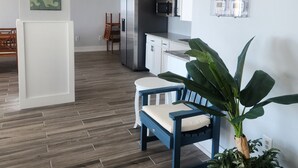 Et moderne køkken med hvide skabe, et rustfrit stål køleskab og en mikrobølgeovn. Der er en blå stol med hynde og en stor potteplante.