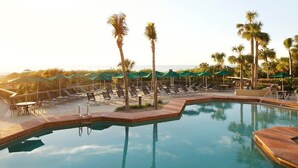 Un área de piscina con sillones de playa y sombrillas, rodeada de palmeras.