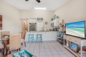 A compact kitchen with white cabinets, a refrigerator, and a microwave. There are two stools and a dining table with chairs. A television is on a stand to the right, displaying a beach scene.