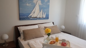 Un lit bien fait, avec du linge de lit blanc, un plateau contenant des articles pour le petit-déjeuner et un tableau représentant des voiliers accroché au mur.