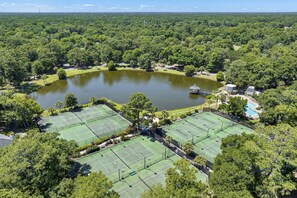 Un complexe de courts de tennis entouré d’une forêt dense, d’un étang et d’un petit pavillon.