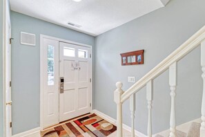 A hallway with a white door, a patterned rug, a small mirror, and a staircase with white balusters.