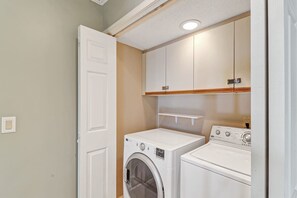 Une buanderie équipée d’un lave-linge et d’un sèche-linge à chargement frontal, avec des étagères en bois au-dessus et une porte blanche.