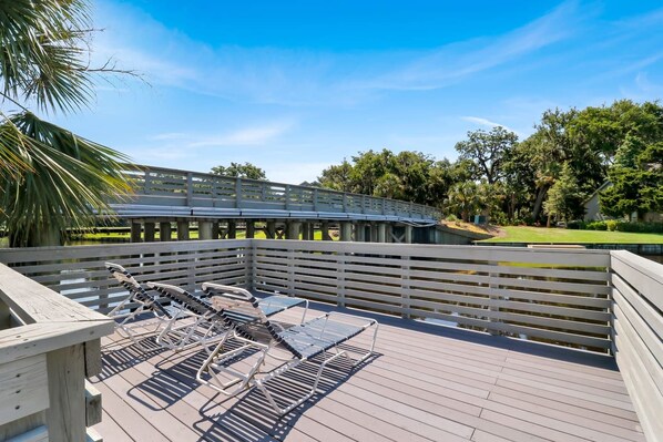A wooden deck with lounge chairs and a view of a park with a bridge and trees.
