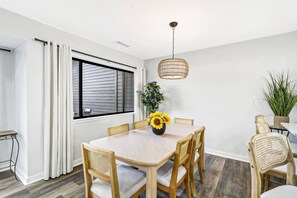 Un coin repas comprenant une table ronde, six chaises, un luminaire suspendu et une fenêtre avec des stores.