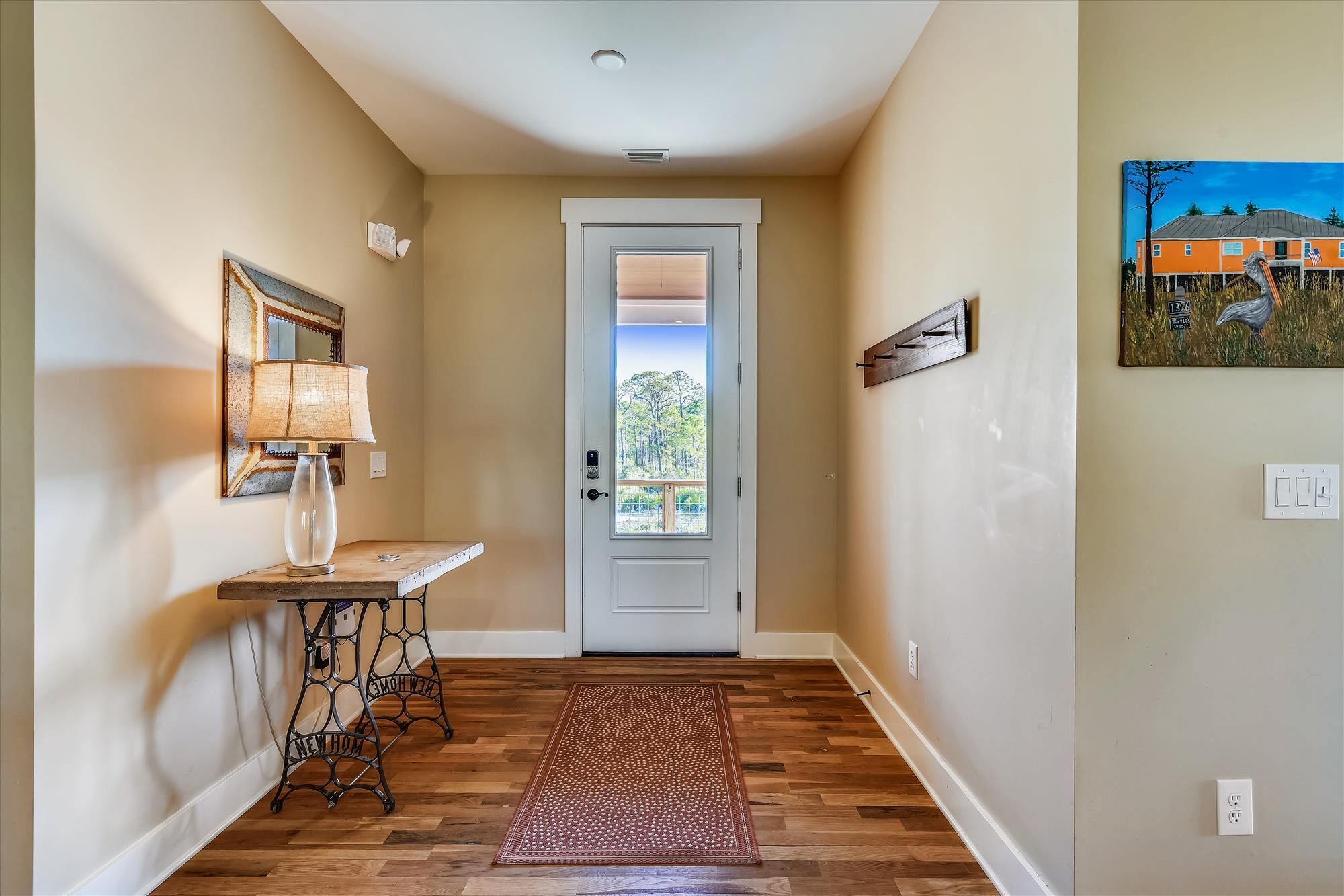 A hallway with a wooden floor, a door with a view to the outside, a lamp, and a painting on the wall.