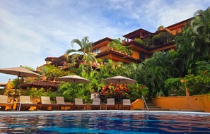 A pool area with lounge chairs and umbrellas, surrounded by lush greenery and a multi-story building with balconies. - Amazing views of La Ropa Bay 3 BR condo (Zihuatanejo)
