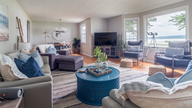 A spacious living room with a large TV, comfortable seating, a dining area, and a view of the outdoors. - Lakefront Renovated 4BR w/ Dock, Kayaks, Walk to Bemus Point (Bemus Point)