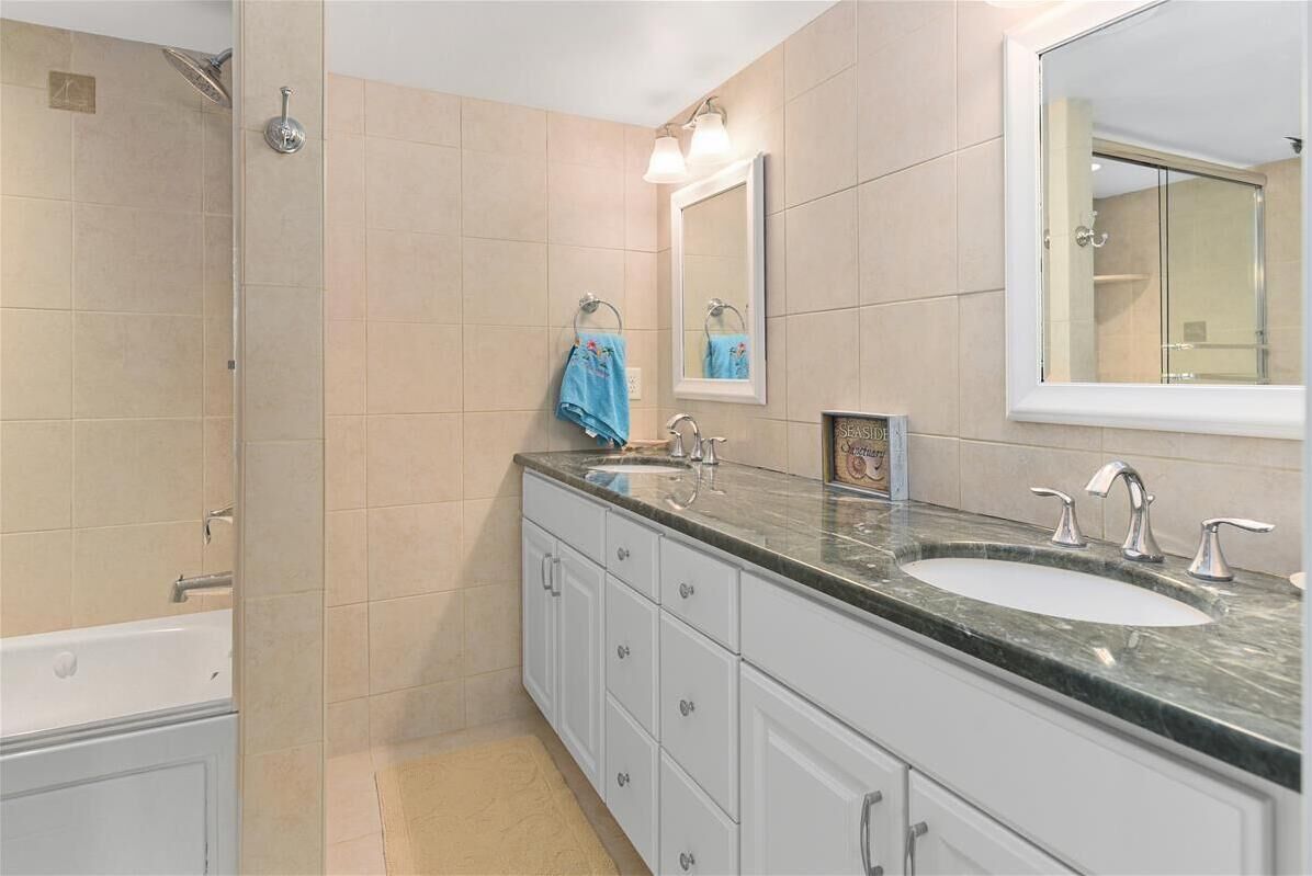Une salle de bain moderne avec des carreaux de couleur beige, un grand miroir, une baignoire et un meuble-lavabo doté d’un évier et de deux robinets.