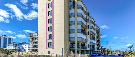 A multi-story hotel with balconies, located near a beach with a wooden fence and a water tower in the background.