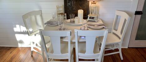 A dining area with a white table set for four, surrounded by white chairs. A candle and a table lamp are on the table.