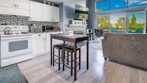 Een moderne keuken met witte kasten, een magnetron en een fornuis. Er is een eethoek met een tafel en stoelen, en een groot raam met uitzicht op bomen en een auto.