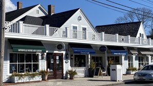 A two-story building with a white facade, a balcony, and storefronts including 'Osterville House and Garden' and 'Coastal Style'. - Osterville Gem with Central A/C and Walking Distance to Joshua’s Pond! (Osterville)