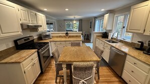 A kitchen with white cabinets, a granite countertop, a central island with wicker chairs, a stove, a toaster, and a sink. - Osterville Gem with Central A/C and Walking Distance to Joshua’s Pond! (Osterville)