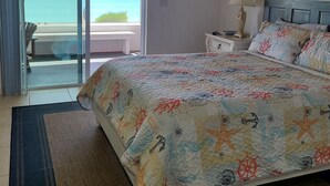 Een slaapkamer met een bed, nachtkastje, lamp, plafondventilator en uitzicht op de oceaan door een schuifdeur.
