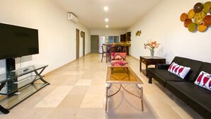 A living room with a TV, sofa, and coffee table. There is a kitchen in the background. - 2 Bedroom Condo a few steps from Médano Beach (Los Cabos)